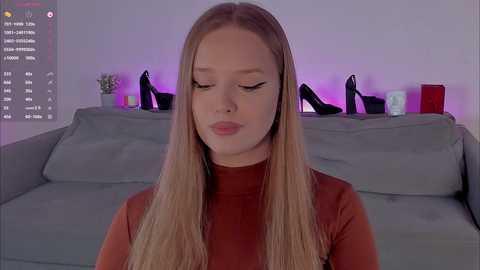 Video of a blonde woman with long hair, wearing a red turtleneck, standing in a modern living room with gray sofa, black high heels, and purple lighting.