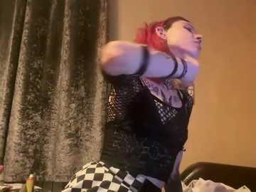 Video of a red-haired woman with pale skin in a black fishnet top, black choker, and checkered pants, striking a pose against a beige wall with dark curtains.