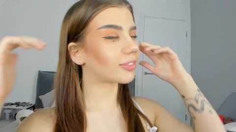 Media: Video of a young woman with light skin and straight brown hair, applying makeup in a modern, minimalist bedroom. She has defined eyebrows and a slight smile, wearing a white tank top.