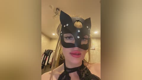 Video of a blonde woman in a black cat mask with silver studs, wearing a black lace choker, in a beige room with a ceiling fan.