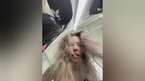 Video of a young woman with messy blonde hair, wearing a black bra, standing in a dimly lit room with hanging clothes and a ceiling fan.