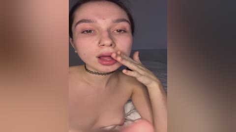 A video of a topless, fair-skinned woman with brown hair, wearing a black choker, licking her fingers in a dimly lit room.