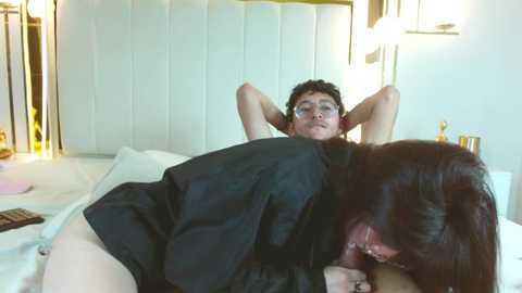 Video of a young man with curly hair and glasses, lying on a bed, wearing a black shirt. A woman with long black hair, wearing a black shirt, is performing oral sex on him. The room is softly lit, with white walls and modern decor.
