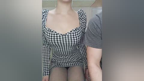 Media: Video of a fair-skinned woman wearing a black-and-white houndstooth dress, sitting on a man's lap. The man, partially visible, wears a grey t-shirt. Background shows a bathroom with a tiled wall.