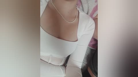 Media: A close-up video of a woman with fair skin, wearing a white, off-shoulder top with a pearl necklace. She has a medium build and is sitting on a striped, patterned cushion.