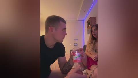 Media: A video shows a young man with short hair and a black t-shirt, holding a soda bottle, looking at a young woman in a red dress, sitting on a bed in a dimly lit room with blue LED lights.