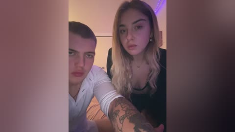 Media: Video of two young people with light skin, one with short hair, the other with long blonde hair, in a dimly lit room, holding hands, looking serious.