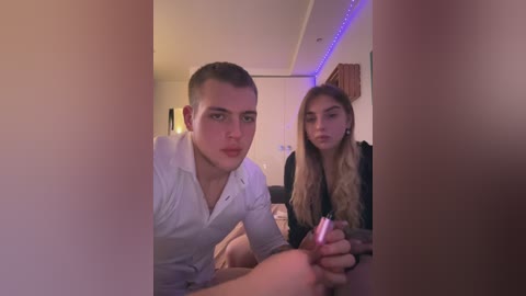 Media: Video of two young Caucasian men, one with short hair and a white shirt, holding a pink lighter, the other with long hair in a black shirt, sitting indoors with dim lighting.