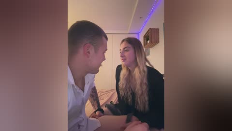 Media: Video of a blonde woman in a black top and a tattooed man in a white shirt, sitting on a bed in a dimly lit, cozy bedroom with purple LED lights.