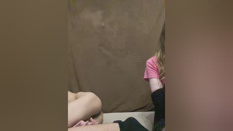 Video of a person in a pink shirt sitting on a beige couch, partially obscured by a brown curtain. The setting appears to be a dimly lit room with neutral tones.