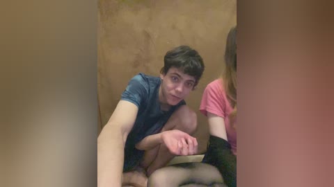 Video of a young man with curly dark hair, wearing a blue t-shirt, crouched in a dimly lit, beige-walled room, with a partially blurred woman in pink beside him.