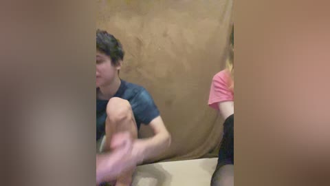 A blurry video shows a young person with short, dark hair wearing a dark t-shirt, sitting in a beige tent, holding a large, pale-colored object. Another person with blonde hair, wearing a pink shirt, is partially visible in the background.