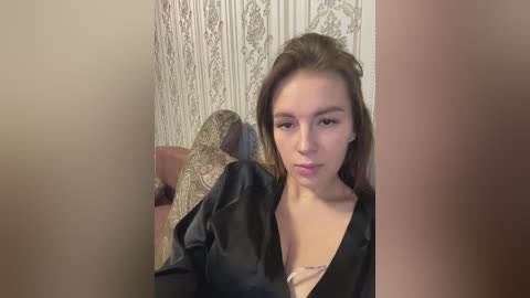 A video of a young Caucasian woman with light skin and straight brown hair, wearing a black satin robe, sitting on a patterned chair in a room with ornate, cream-colored wallpaper.
