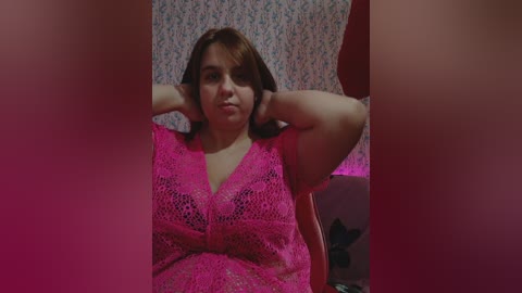 Video of a plus-sized woman with light skin and brown hair, wearing a bright pink crochet top, sitting with arms behind her head, against a floral-patterned wall.