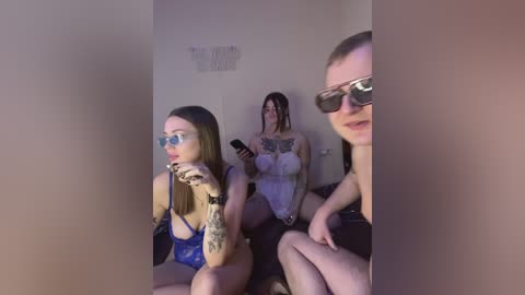 A video captures three women seated in a dimly lit room, each wearing revealing clothing. One woman with tattoos, glasses, and blue lingerie, another in a sheer white top, and the third in a black top.
