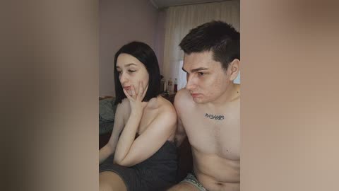 Video of a young couple sitting close on a bed, both shirtless; woman with fair skin and shoulder-length black hair, man with short dark hair, looking at each other.
