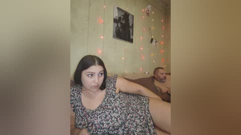 Video of a young woman with shoulder-length brown hair, wearing a floral dress, looking upset, sitting beside a man with a beard, in a dimly lit room with string lights and a framed photo on the wall.