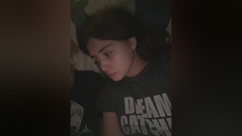 Media: Video of a young woman with medium skin tone and curly black hair, lying on a dark bed with a \"DEAD CAT\" t-shirt, dim lighting, and blurry background.