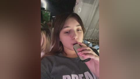 Media: A video captures a young Asian woman with long, straight hair drinking from a can, wearing a gray \"BREND\" t-shirt, in a dimly lit, cluttered room with a metal door.