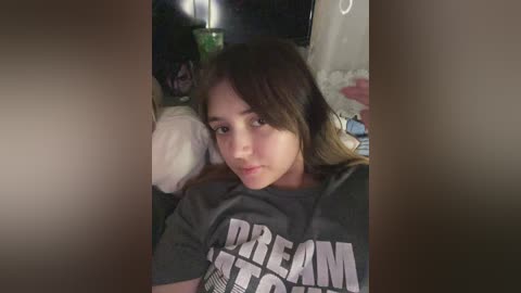 Media: Video of a young woman with long brown hair, wearing a gray \"Dream\" t-shirt, lying on a bed with a green blanket, dimly lit room in the background.