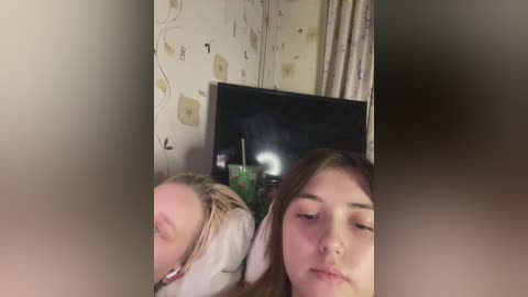 Media: A video of two young girls with light skin and long brown hair, lying side by side, eyes closed, in a cozy bedroom with floral wallpaper and a TV screen in the background.