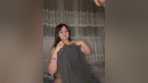 Media: Video of a young woman with light skin and straight brown hair, holding a dark towel in front of a lace curtain backdrop.