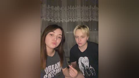 Media: Video of two young women, one with long dark hair, the other with short blonde hair, both wearing dark t-shirts. They are kissing, indoors with a lace curtain backdrop.