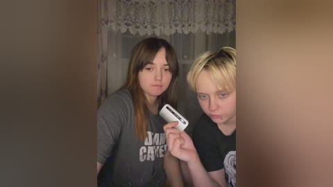 Media: Video of two young women, one brunette and one blonde, wearing casual T-shirts, standing in a dimly lit room with lace curtains and a beige wall. The blonde holds a camera.