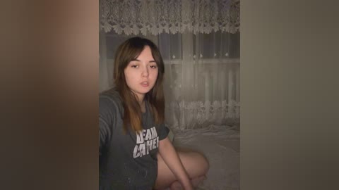 Media: A video of a young Asian woman with long brown hair, wearing a gray \"Lena Dunham\" t-shirt, sitting on a bed with white lace curtains and a soft, dimly lit room.