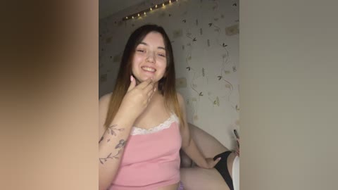 Media: Video of a smiling Latina woman with long brown hair, wearing a pink tank top and black panties, sitting on a bed in a dimly lit room with butterfly-themed wallpaper and fairy lights.