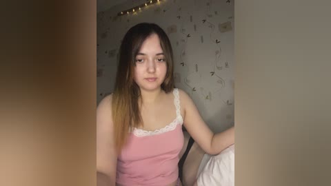 Media: A video of a young woman with long, dark hair and light skin, wearing a pink camisole with lace trim, sitting on a bed in a dimly lit room with patterned wallpaper and fairy lights.