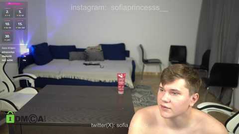 Media: Video of a young, shirtless man with short brown hair, sitting at a dark wooden table in a modern, minimalist living room with a blue sofa, chairs, and a TV.