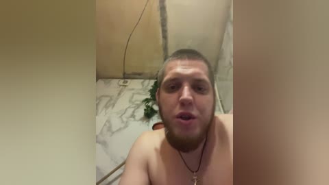 A video of a shirtless, fair-skinned, bearded man with short hair, wearing a necklace, standing in a dimly lit room with marble walls and a green plant.