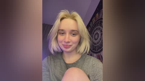 Video of a young woman with shoulder-length, platinum blonde hair, fair skin, and a light gray shirt. Background includes a dark tapestry with circular patterns and a curtain.