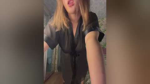 Video of a fair-skinned woman with blonde hair, wearing a black sheer blouse, standing in a dimly lit room with floral wallpaper.