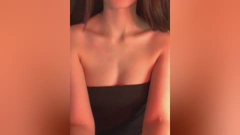 Video of a light-skinned woman with long brown hair, wearing a black strapless dress, against a warm, orange background, highlighting her bare shoulders and collarbone.
