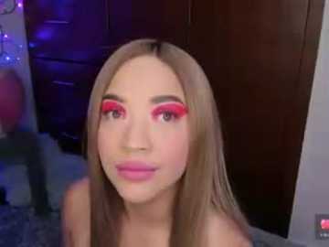 Video of a light-skinned woman with long, straight, light brown hair, wearing bold pink and red eyeshadow, smiling slightly, indoors, blurred background.