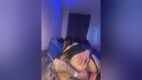 Video of a woman in black lingerie, lying on a bed with blue and purple lighting, tattooed arm, and another person's legs visible.