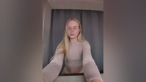 A video of a young, fair-skinned, blonde woman with long hair, wearing a sheer, long-sleeved top, standing in a mirror hallway.