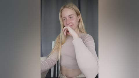 Video of a blonde Caucasian woman with long hair, wearing a light pink long-sleeve crop top, smiling while touching her hair, in a neutral-toned room with gray walls.