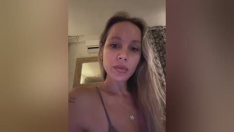 Media: Video of a fair-skinned, young woman with long, straight, light brown hair, wearing a grey tank top, standing indoors near a window with floral curtains.