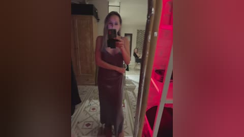 Media: Video of a woman in a brown, sleeveless dress taking a mirror selfie in a dimly lit bedroom with red lighting.