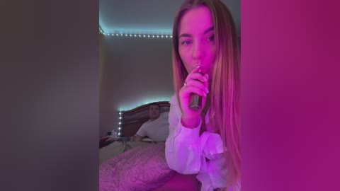 Media: Video of a young woman with long blonde hair, wearing a white blouse, holding a green vape pen, in a bedroom with pink and blue LED lights, wooden bed, and textured blanket.