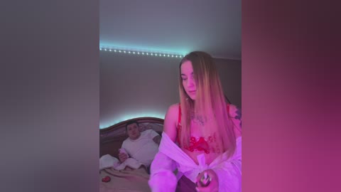 Media: Video of a blonde woman in red lingerie, standing in a dimly lit bedroom with neon lights, next to a man in a white shirt.