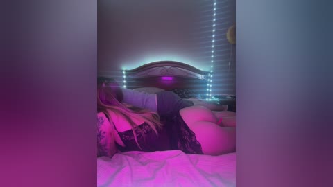 Media: Video of a woman lying on a bed with pink and blue lights, wearing black lace lingerie, showing her bare buttocks, in a dimly lit bedroom.