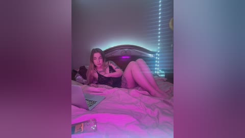 Media: Video of a young woman with long hair, wearing lingerie, lying on a bed with a laptop, under dim, pinkish-blue lighting.