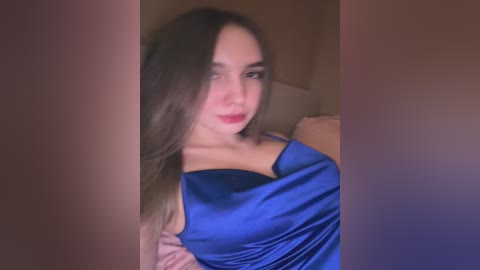 Media: A video of a young woman with fair skin and long brown hair, wearing a bright blue satin dress, smiling seductively. The background is dimly lit, with blurred edges, giving a sensual, intimate atmosphere.