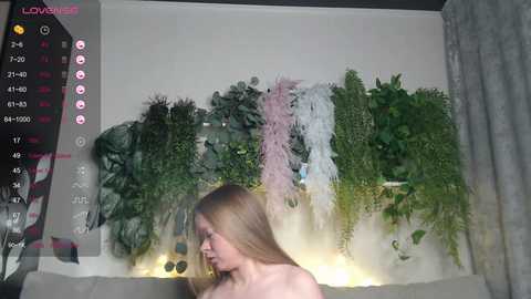 A video of a nude woman with long blonde hair, standing in front of a lush green wall of hanging plants. The scene includes a digital camera interface overlay.