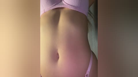 Media: Video of a close-up of a woman's midriff, wearing light pink lingerie, with a soft, warm light creating a gentle glow on her skin. The background is blurred and beige.