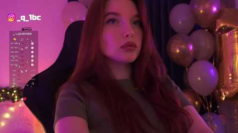 A video of a fair-skinned woman with long, auburn hair, wearing a dark top, seated in a chair amidst a room filled with purple and gold balloons and string lights.